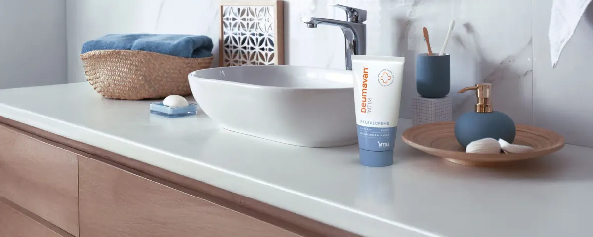 A bathroom counter with a washbasin, a tube of the intimate care cream, a blue cup with a toothbrush, a blue dispenser on a wooden tray and a basket with a rolled blue towel.