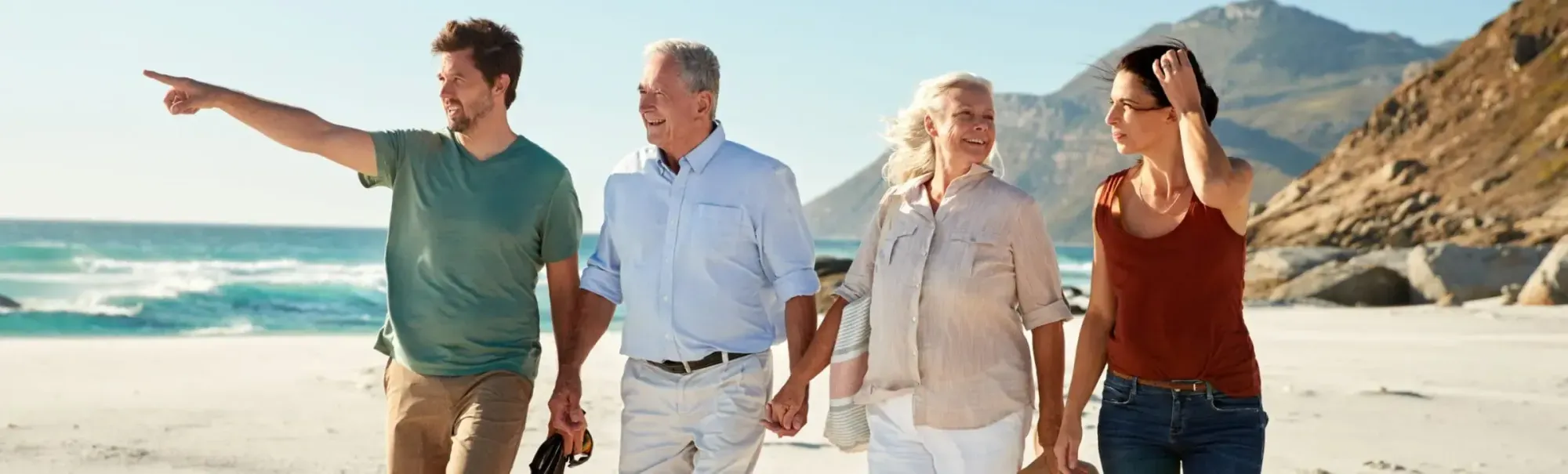 Four adults of different ages walking hand in hand along a sandy beach by the sea.