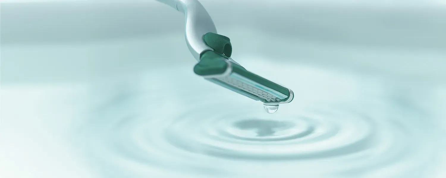 The head of a razor touching still, pale turquoise water, with a drop falling from it and rings spreading out.