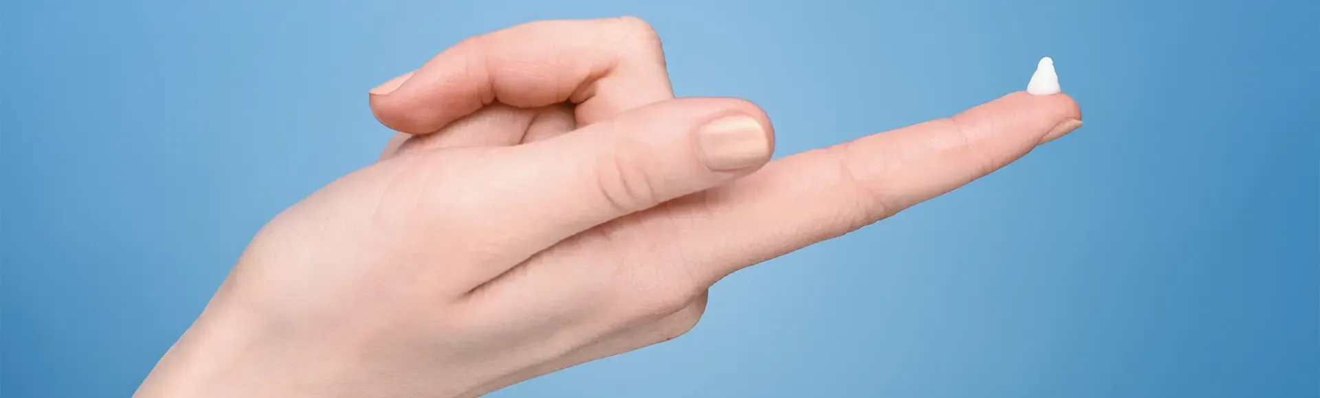 A hand against a blue background holding out its index finger, with a small dab of white ointment on the fingertip.