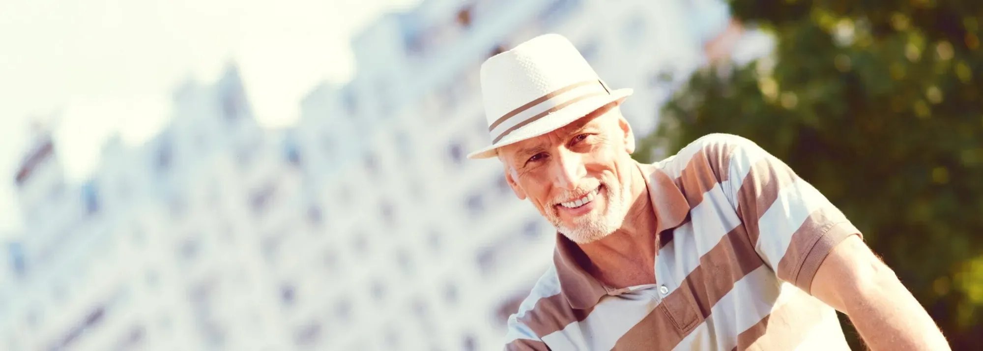 An older man in a straw hat and a striped polo shirt smiling outdoors, high-rise buildings blurred behind him.
