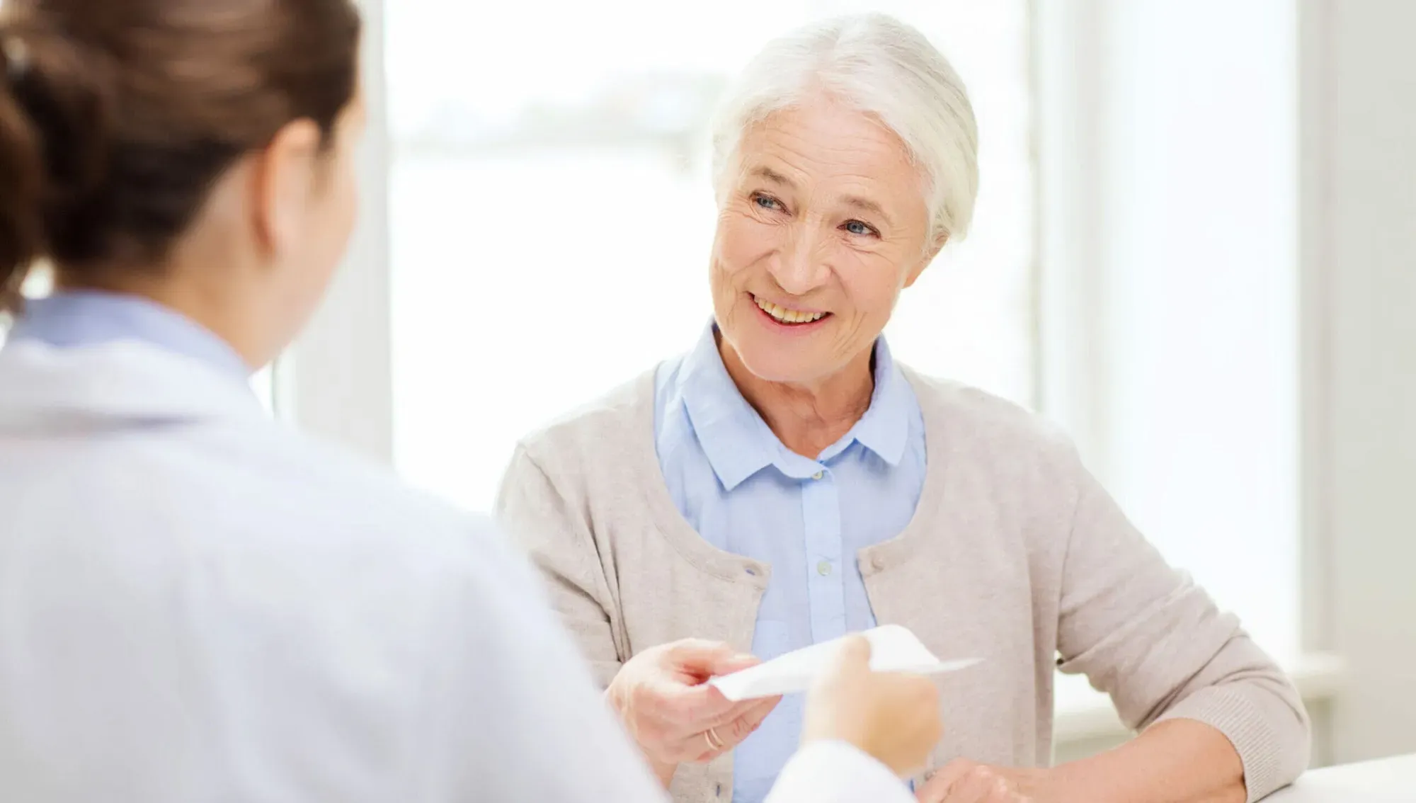 An older woman smiling across a desk at a doctor in a white coat, who is handing her a sheet of paper.