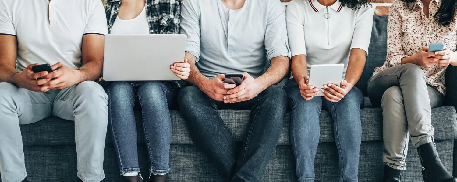 Five people sitting side by side on a grey sofa, looking at their phones, a laptop and a tablet.