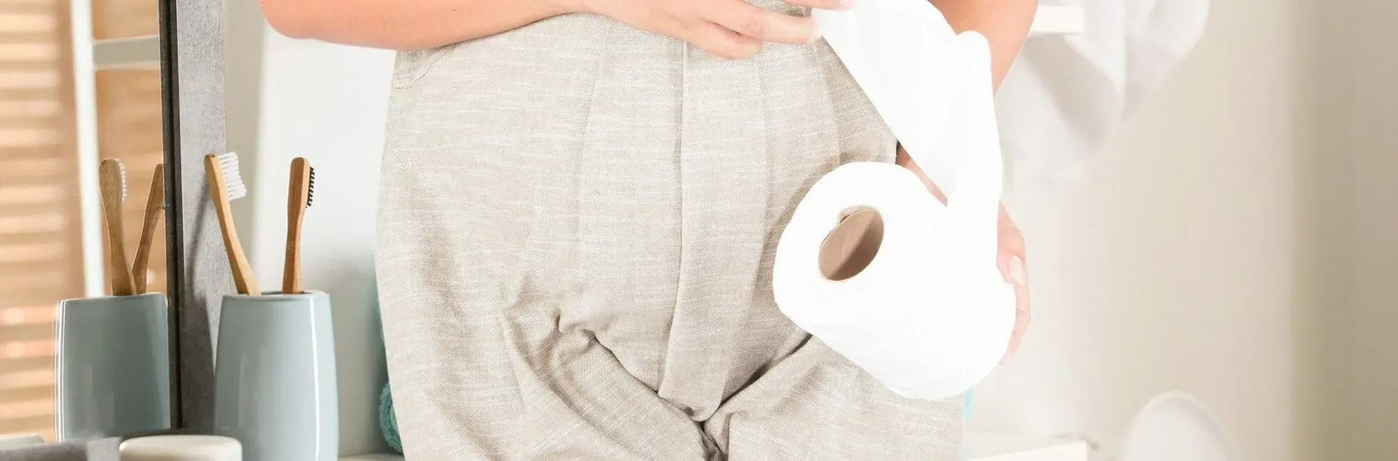 A pregnant woman in linen dungarees in a bathroom, one hand on her belly, holding a roll of toilet paper.