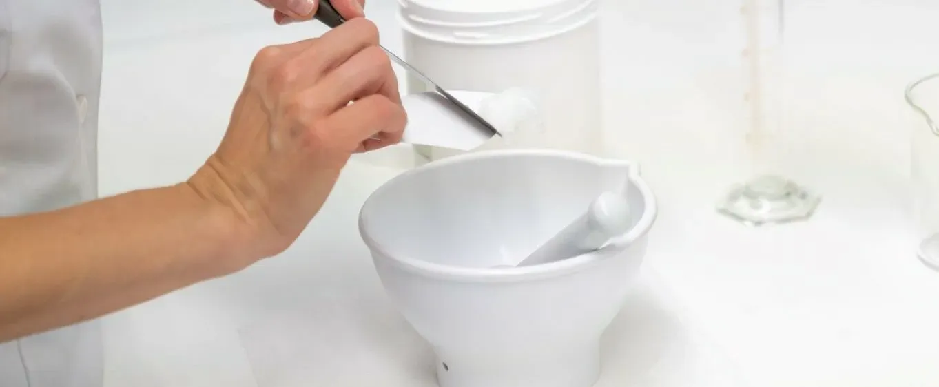 A hand lifting white petroleum jelly out of a jar with a spatula, above a white mortar and pestle; laboratory glassware in the background.