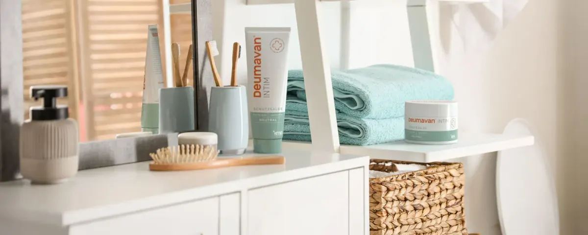 A white bathroom shelf with a tube and a tin of the protective ointment, toothbrushes in cups, a soap dispenser, a hairbrush and folded turquoise towels.