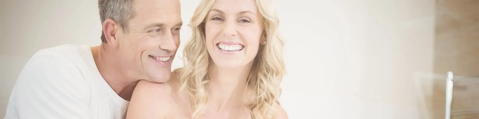 A smiling man and woman close together in a bright room, his chin resting on her shoulder.
