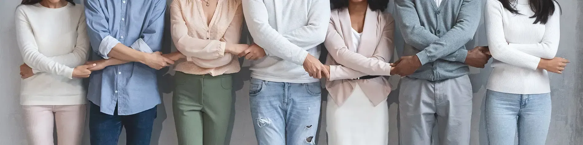 A row of women and men in casual clothes standing side by side against a grey wall with their arms linked.