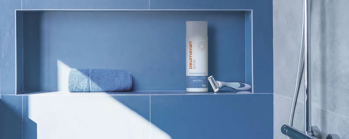 A blue-tiled shower niche with a bottle of the washing lotion Neutral, a razor and a rolled blue towel.