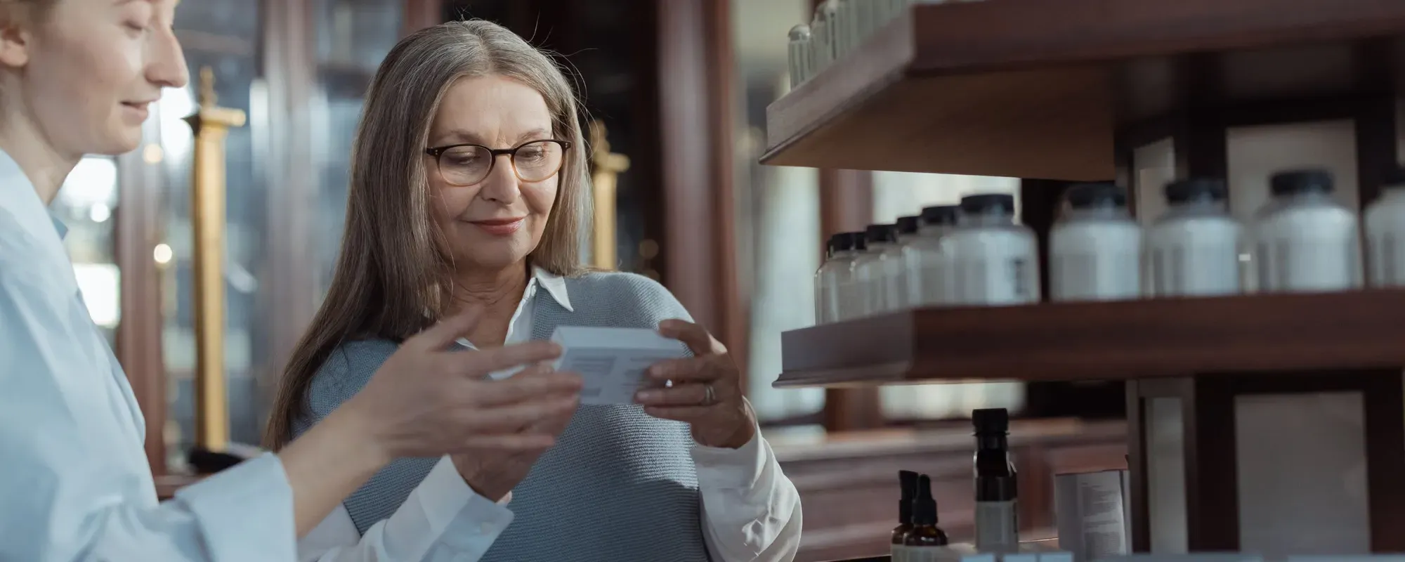 Dans une pharmacie, une femme aux cheveux gris et aux lunettes regarde une petite boîte que lui tend une femme en chemise bleu clair, devant des étagères en bois garnies de flacons blancs.